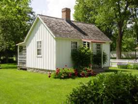 A small, white two-room cottage with a white picket fence and a flower bed.