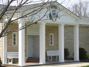 Four white columns and a white portico on a buff-colored brick building with a slate roof.