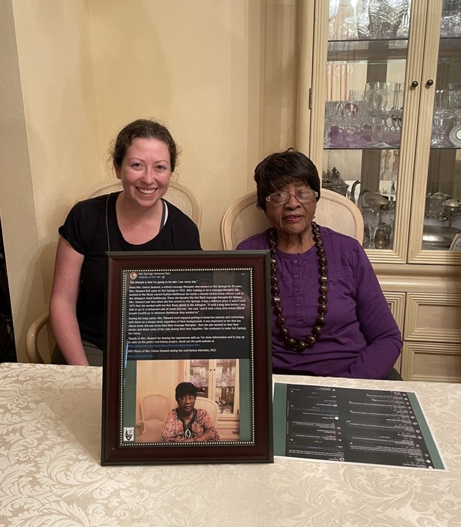 Two women sit behind a table with a framed photo on the table