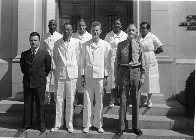 Eight people stand in front of a building, 5 men and 3 women. Two men are dressed in suits and the other six are in all white uniforms