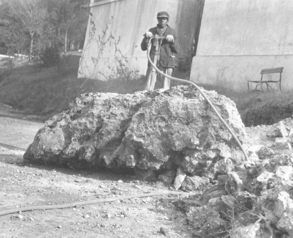 Man in dark jacket and hat holds jackhammer while standing on large rock