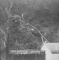 black and white 1867 photo of a wooden flume coming down the hillside to a collecting tank built to the left of the bathhouse building