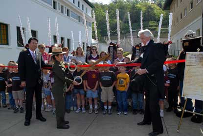 Hanamaki, Japan, mayor, Superintendent Josie Fernandez with large scissors, re-enactor President Andrew Jackson, and Smokey Bear in front of red ribbon, young school children behind ribbon and Deer Dancers behind children.