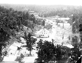 black and white photo from stereograph; looking south on the hot springs valley with mountain on left and small wooden buildings along either side of a dirt road; some foundations visible of buildings burned during the Civil War