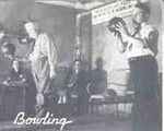 Old black and white photo of two men bowling; each is holding ball ready to roll it down the lane; one man sits in the background