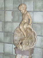 Light gray glaze colored cherub sitting on a shell shape surrounding a fish head that was a fountain. Surrounding tile is a blue green color