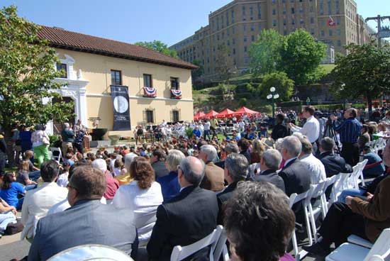 Shot from back of crowd, looking toward front of park administration building. Mint director is speaking from podium to left of fountain. Building front has coin banner and bunting hanging from 2nd story windows.