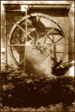 sepia tone photo of end of stone building with metal water wheel attached to it