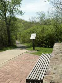 beginning of sunset trail at West Mountain Summit overlook, with stone wall and bench on lower right, gravel trail in middle with brown trailhead sign and trees in background