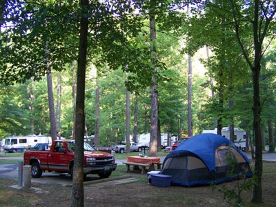 Blue dome tent in foreground with red pickup truck parked at campsite. In background you can see RV campers. All with trees surrounding.