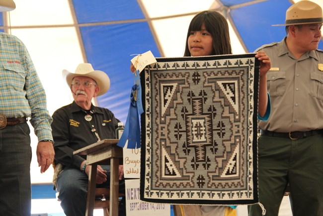 A young Navajo Weaver is holding up her rug during an auction.