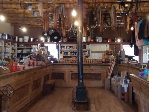 Interior of a historic trading post general store. There are wooden counters on three sides of the room with a cast iron stove in the center. a variety of foodstuffs and mercantile items are behind the counters.