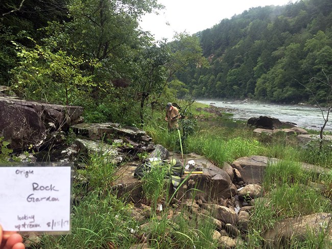 Monitoring equipment laid out on a rock, and a person with a clipboard beyond, in a grassy area beside a river. In the corner of the frame a note identifies the site as the "Rock Garden."