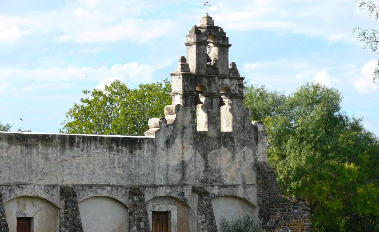 old mission building with a bell tower and forest in the background