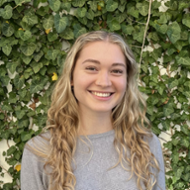 A woman stands smiling in front of a wall covered with ivy.