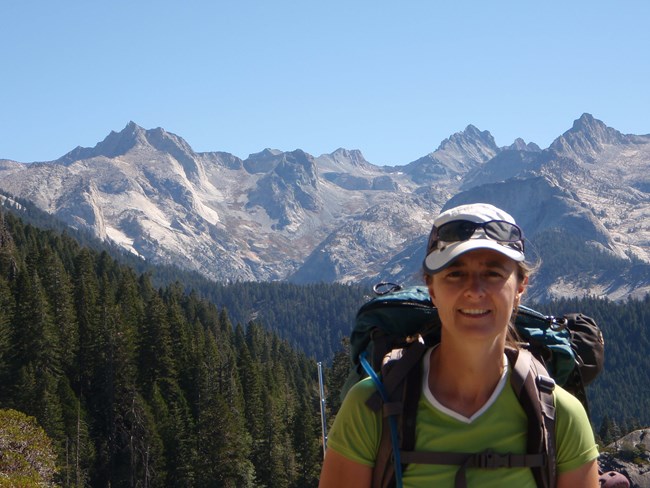 Woman wearing backpack, and faces camera smiling with view of mountain range behind her.