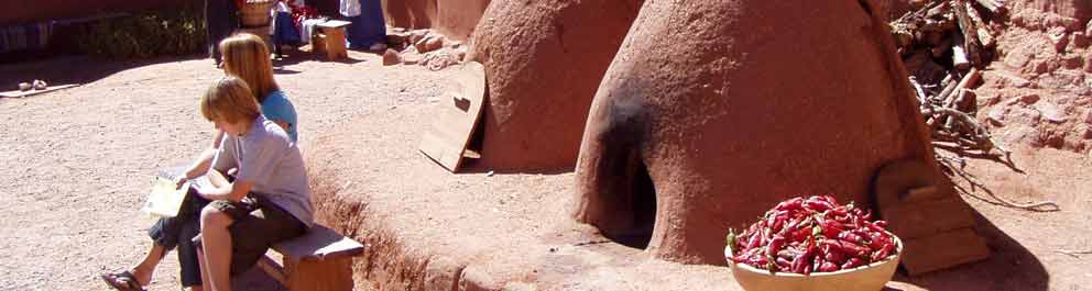Two children sit in bright sunshine next to earthen horno and chili bowl