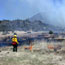 color photograph of firefighter watching burning grass with volcano obscured by smoke in background
