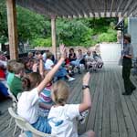 Visitors at an evening program held on the Visitor Center porch raise their hands to respond to the ranger.