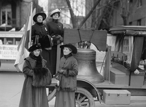 Historic black and white photo of a replica of the Liberty Bell on the back of a truck, surrounded by women dressed in long dresses and hats.