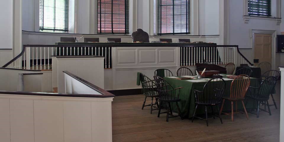 Color photo of courtroom with six chairs on dais, raised above a wooden jury box, and one table surrounded by Windsor chairs.