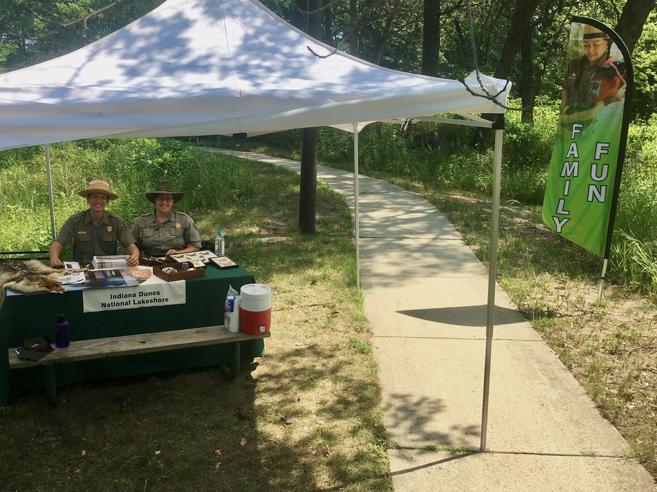 Two park rangers sit at a table under a tent outside.