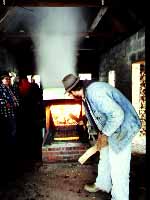 man putting a log inside a roaring fire box with a pan onto with steam rising above it