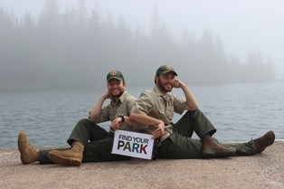 Two visitor services volunteers pose on the ground in front of Lake Superior for a photograph with a Find Your Park sign.