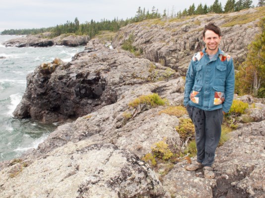 A man smiles while standing on rocky land with the shoreline to his left