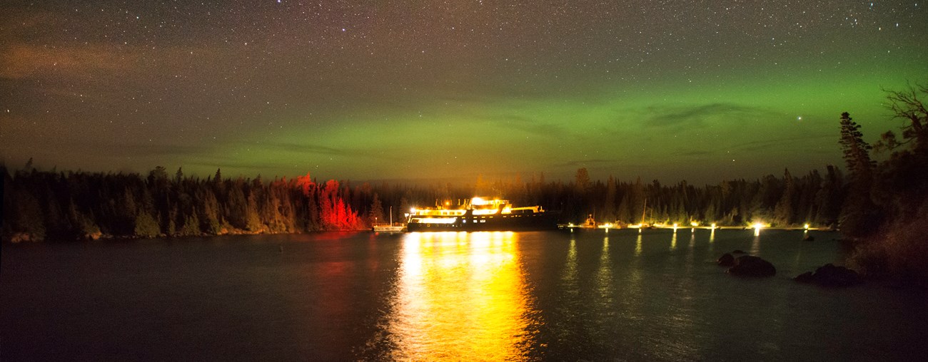 Ranger III Ferry at dock at night in Rock Harbor. Lights reflect of the of the dark water. Green waves of the aurora borealis dance above the treeline against a star-filled sky.