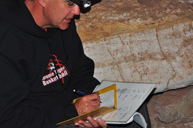 A teacher wearing a helmet and headlamp writes in a record book inside a cave passage.
