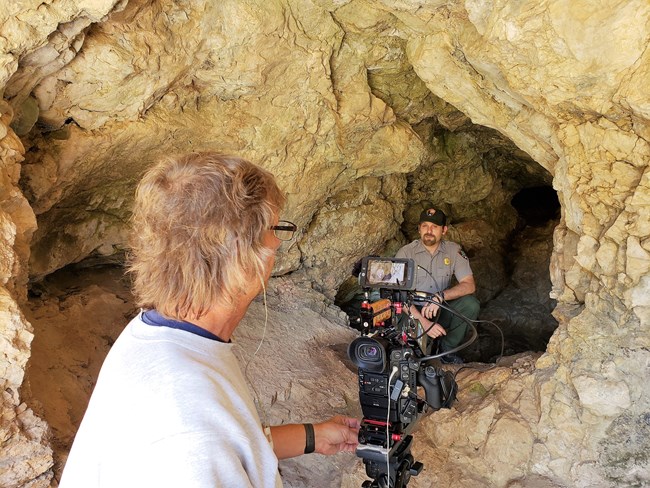 A park ranger is being filmed while kneeling inside a cavern surrounded by tan-colored rocks.