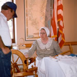 A park ranger in an old-fashioned dress and cap shows a visitor how a spinning wheel works at the Prairie Acadian Cultural Center