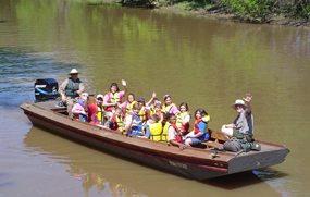 Children and a park ranger wave from a traditional wooden boat during a bayou tour