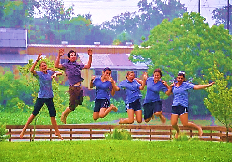 A field with a wooden fence in the distance. 6 people jumping in their air and smiling.