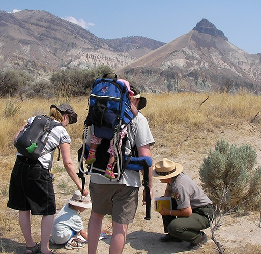A ranger shows a young visitor fossils found in the area while parents look onward. One of the parents has a baby on his back riding in a carrier. Sheep Rock is seen in the distance.