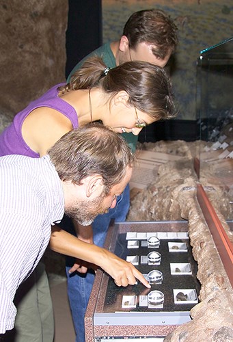 Three adults look at the fossils on display in the drawers at the visitor center. The fossils have magnification lens on them to make them easier to see the details. One points to a center fossil.