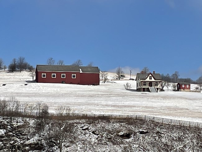 A red barn building and a farmhouse on a hillside with snow.
