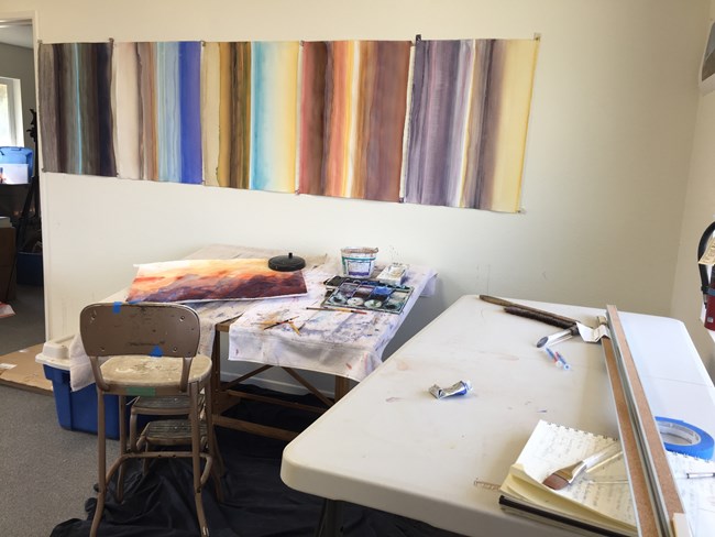Indoor artist in residence studio.  In the foreground a white table with various art supplies.  In the back ground a painting of mountains dries on a desk and gradient artwork hangs on the wall.
