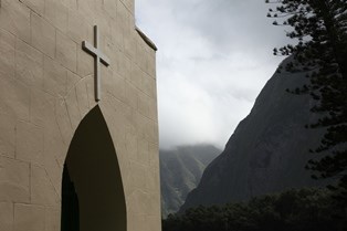 A close up of the entrance way to a church building with a large cross on top of the entrance. In the background tall, green sea cliffs rise towards the sky.