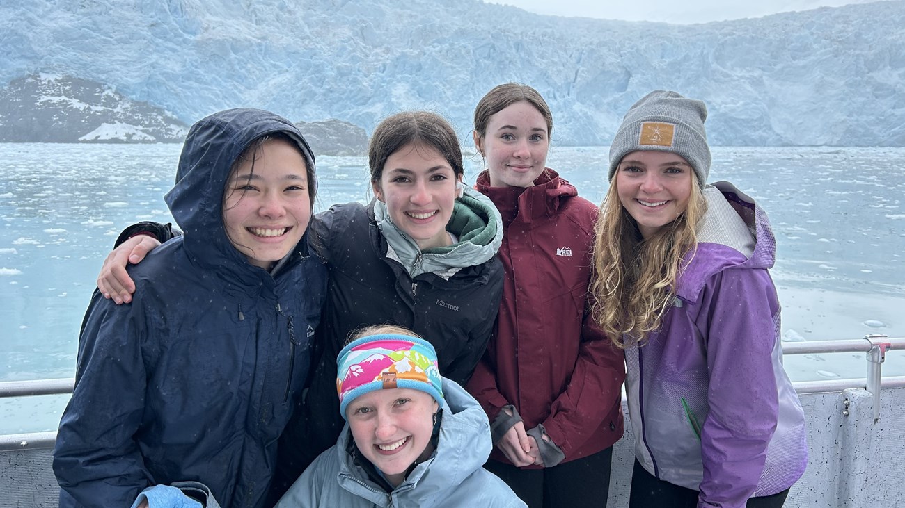 Five youth employees pose together on a boat in front of a large tidewater glacier.