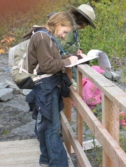 A student stands on a bridge, next to a park ranger, and writes down observations about the plant life of Kenai Fjords.