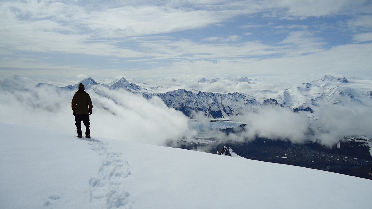 A person standing on a snow looks out towards a bay and snow-covered mountain ranges in the distance