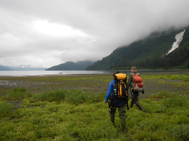 Two people with backpacks walk towards water between green mountains below grey clouds.