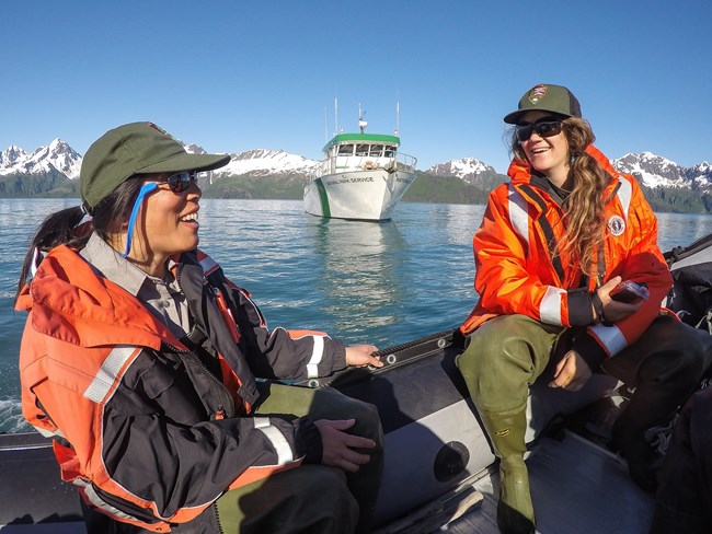 Two rangers with bright orange jackets smile talking while on the water with a boat reading National Park Service beyond.
