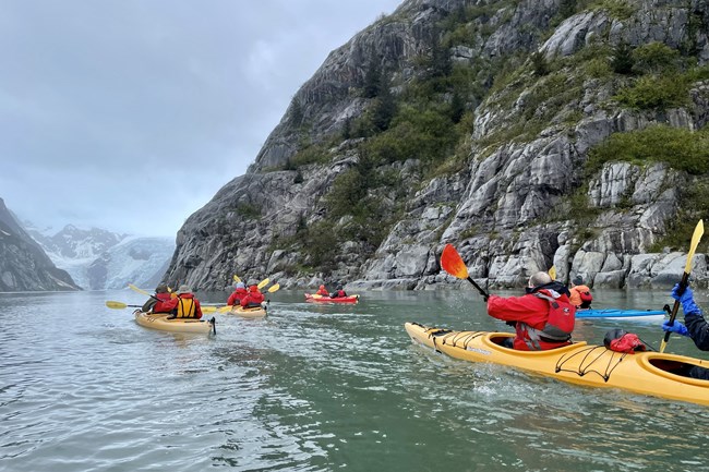 People in multiple kayaks paddle in a fjord toward a glacier.