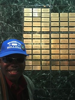 Black man in blue baseball cap and glasses smiles next to a plaque of gold-plated names on a wall.