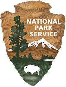 NPS logo inside arrowhead shape. Displays a tall tree, snow-capped mountain, lake, green grass, and a bison. Text: National Park Service.