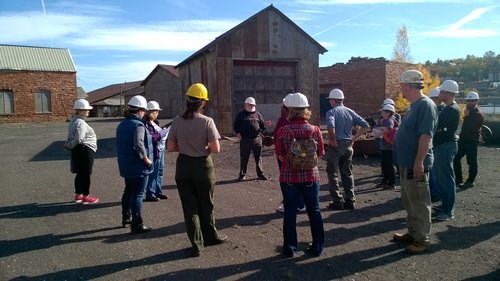 People with hardhats on listen to a speaker at an outdoor industrial site.