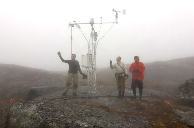 Three people stand on a rocky area in the fog next to a metal tower